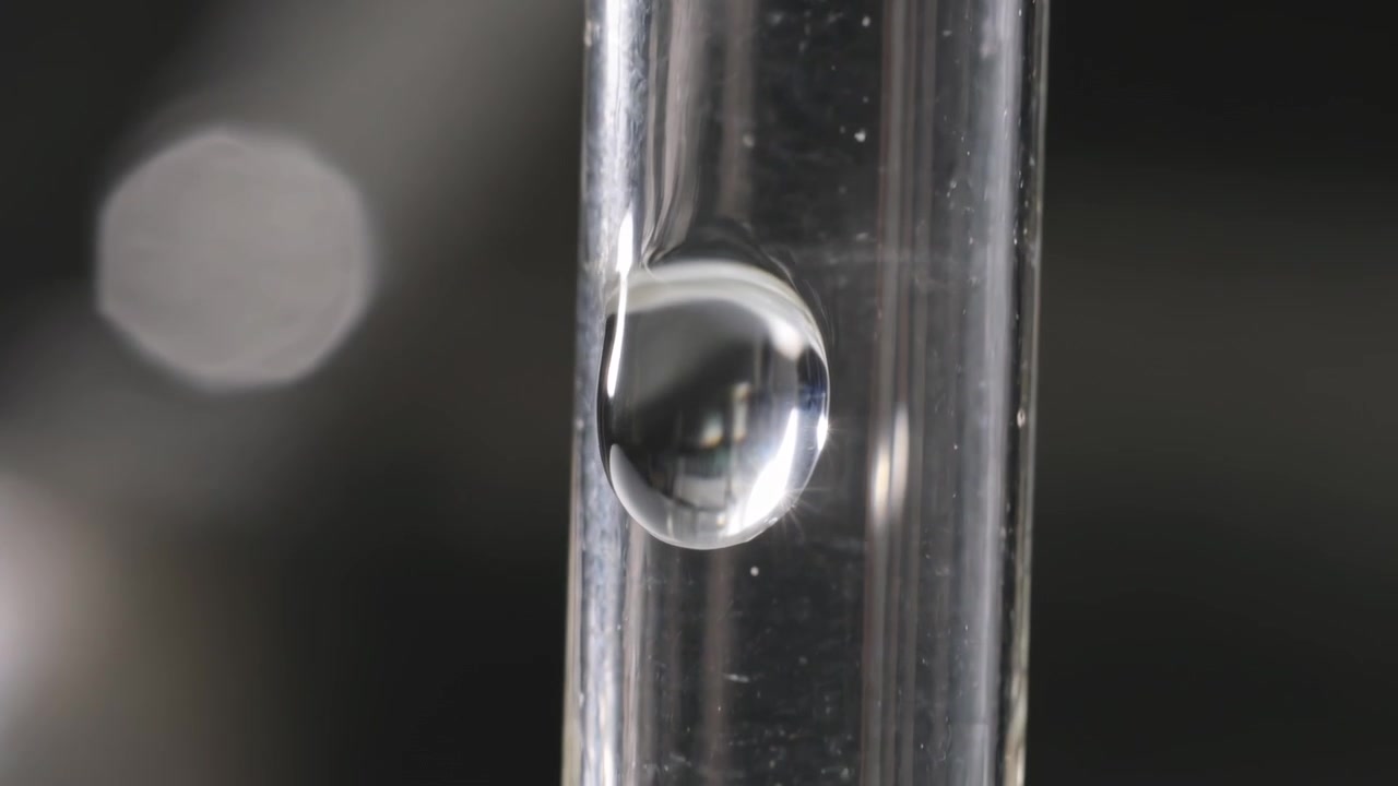 Gravità che attira una singola goccia trasparente che scivola lungo un tubo di vetro verticale con piccole bolle e bokeh