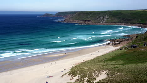 ocean waves along the wide sandy shore at sennen cove in cornwall with surfers and tourists walking on the beach