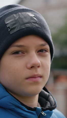 primer plano de un niño con gorra negra y chaqueta azul al aire libre, con expresión tranquila y ligero ceño fruncido, rodeado de vibrantes árboles de otoño y un fondo borroso con casa de techo rojo y vegetación