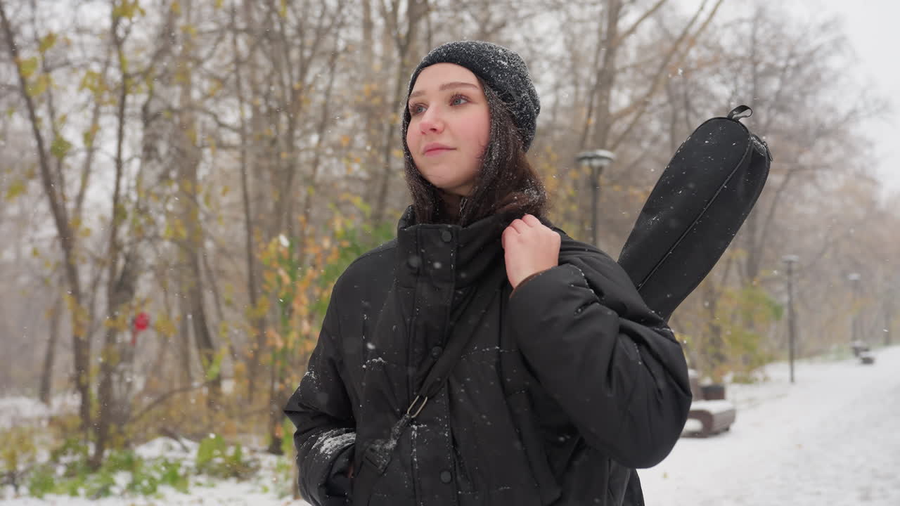 Girl in black hoodie adjusting her guitar in its pack, looking around thoughtfully, with a snow-covered bench in the background amidst a serene snowy landscape