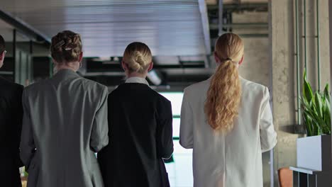 Rear-view-of-a-group-of-confident-businesswomen-in-business-clothes-walking-along-the-corridor-in-a-modern-office.-Confident-girls-in-business-clothes-walk-along-the-office-corridor-together