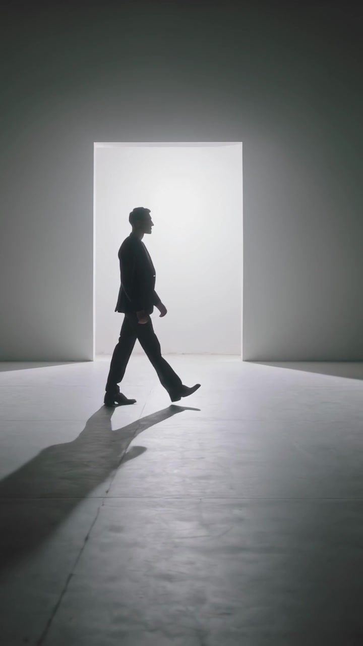 Video verticale: Entrando nell'inquadratura, uomo in abito scuro che cammina verso una porta illuminata in studio, proiettando un'ombra