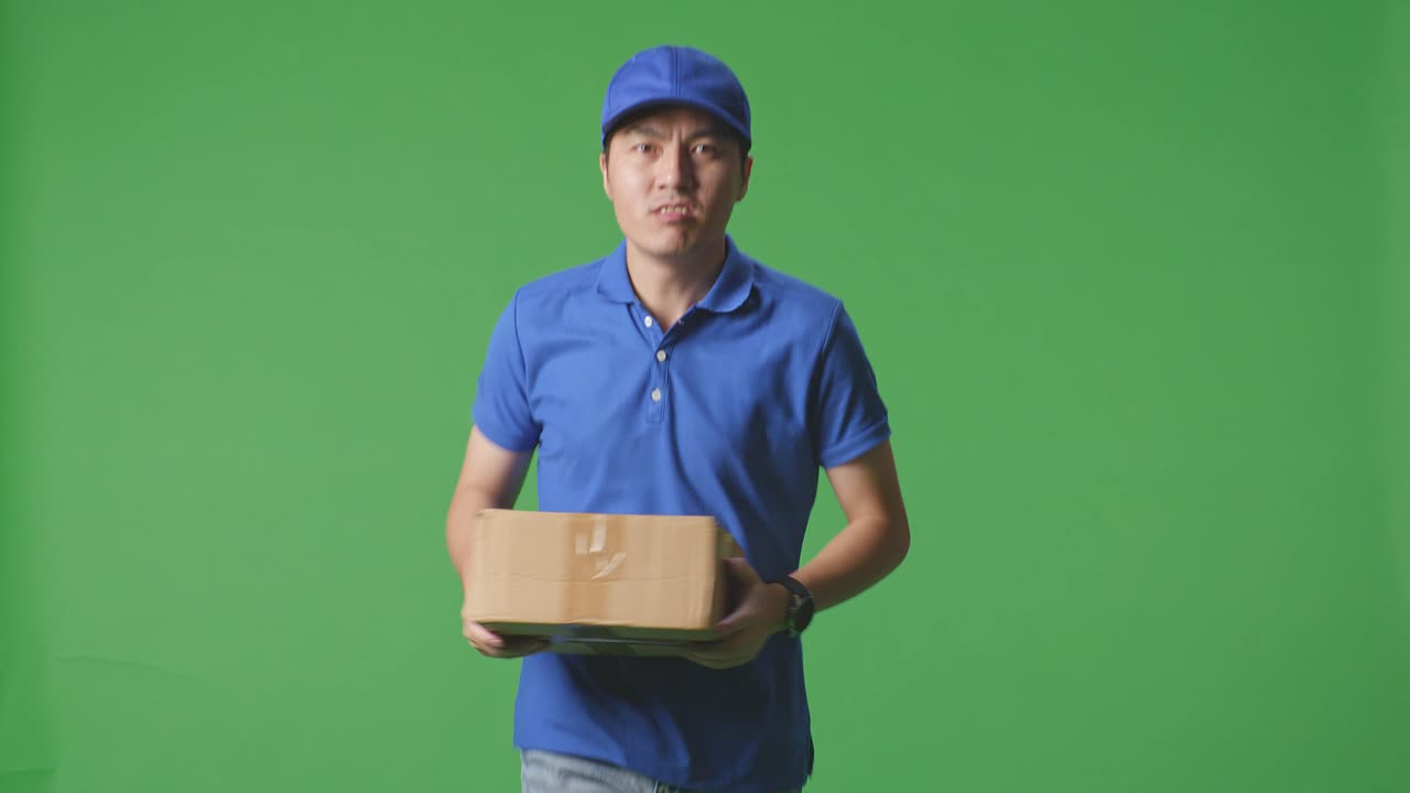 Asian Male Courier In Blue Uniform Running While Delivering A Carton On Green Screen Background In The Studio