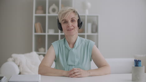 charming middle-aged woman with headphones is greeting by videocall looking at camera sitting in room medium portrait