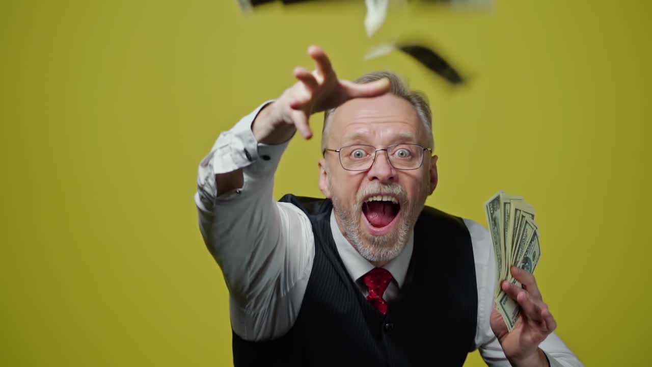 Happy elderly man throwing money. Mature man in glasses with grey hair holding dollar banknotes and throws into the air indoors. Slow motion.