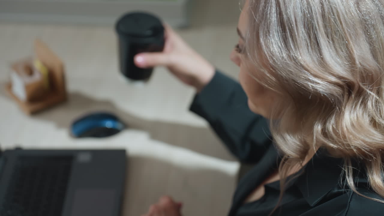 Top down close up of woman with wavy blonde hair reaching for coffee while working at desk with laptop, mouse, and office supplies, briefly pausing workflow in brightly lit modern workspace