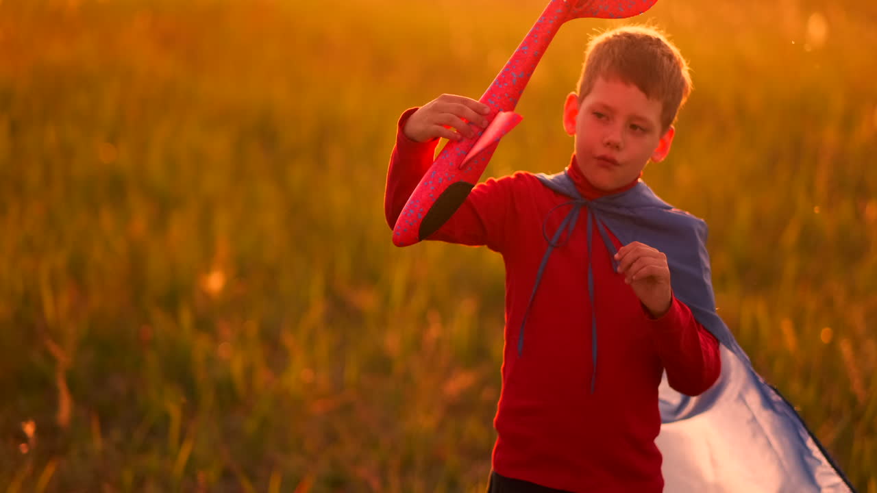 el niño con el disfraz de un superhéroe corriendo en una capa roja riendo al atardecer en el campo de verano representando que él era el piloto del avión jugando con un modelo de avión