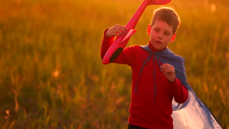 the boy in the costume of a super hero running in a red cloak laughing at sunset in summer field representing that he was the pilot of the plane playing with a model airplane