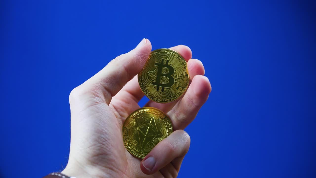 A close-up of a hand holding Ethereum and Bitcoin coins against a blue backdrop