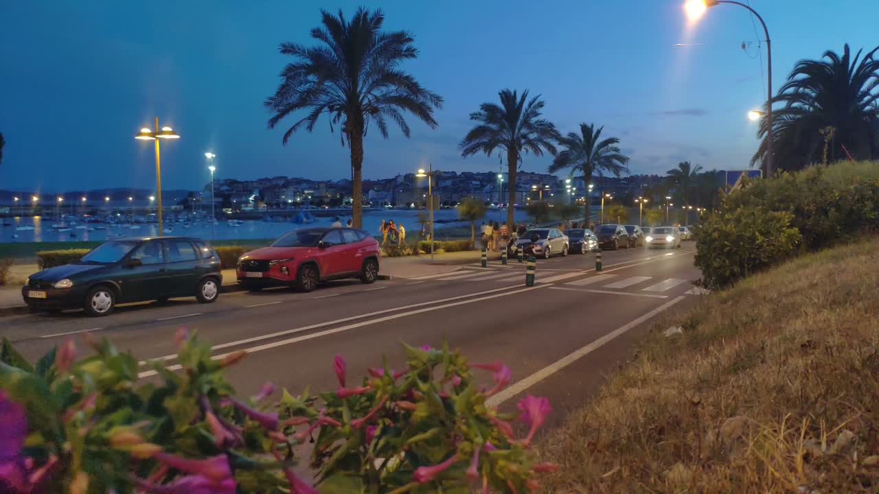people walking along the sidewalk among palm trees, cars circulating on the road with street lamps at sunset, the beach and the port in the background. Panoramic shot. Porto-Novo, Pontevedra, Spain