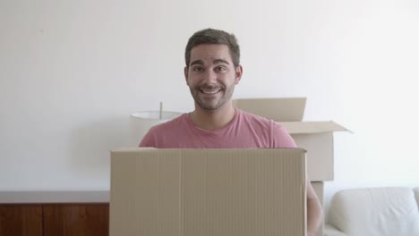 portrait of happy young man standing in the living room of his new house, holding a cardboard box and smiling at the camera