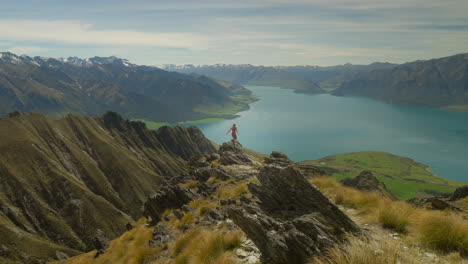 Mujer-Aventurera-Saltando-Desde-Roca-Irregular-En-Paisaje-Alpino-Con-Lago