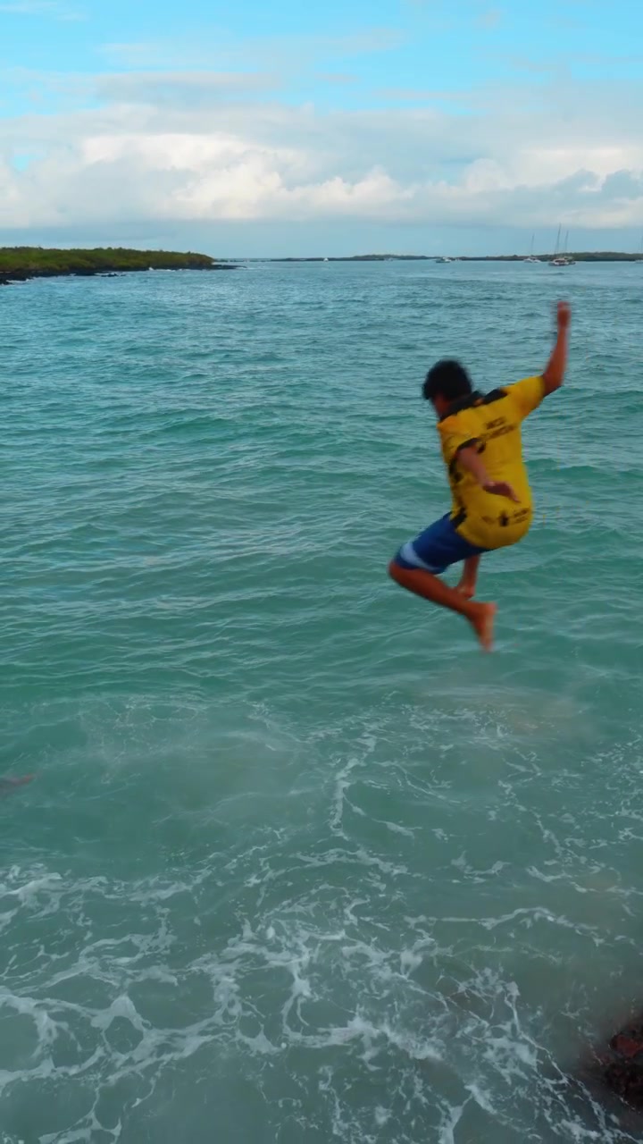 Santo Tomas, Ecuador, 1 maggio 2026: Ragazzo che si tuffa in acqua turchese sulla costa delle Galapagos. Ragazzo con maglietta gialla si tuffa verso l'acqua turchese con schiuma, rocce e barche in lontananza