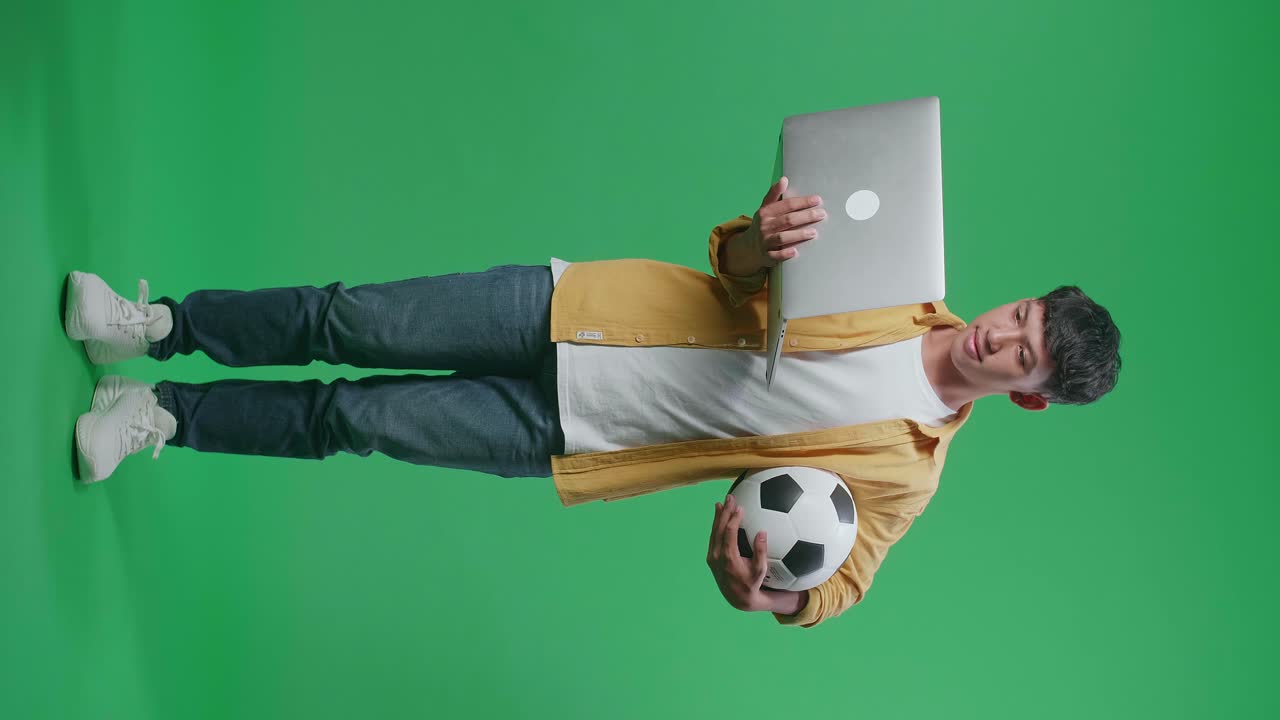 cuerpo lleno de niño asiático con una pelota viendo un partido de fútbol en una computadora portátil en el estudio de pantalla verde