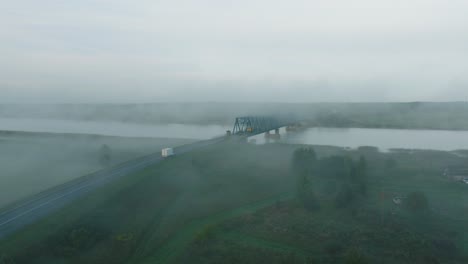 Luftaufnahme-Der-Stahlbrücke-über-Den-Fluss-Lielupe-An-Einem-Sonnigen-Sommermorgen,-Nebel-Steigt-über-Dem-Fluss-Auf,-Fahrende-Autos,-Breite-Drohnenaufnahme,-Die-Sich-Tief-Vorwärts-Bewegt