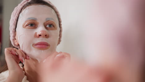 woman relaxes doing beauty treatments using face mask. pretty female with manicure in bathrobe puts on face mask standing in bathroom slow motion