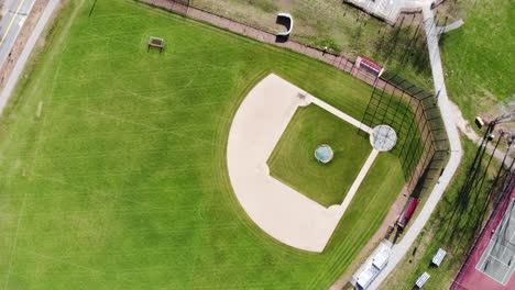 a daytime drone video with a birds-eye perspective over a vacant baseball field in the summer, showing tennis courts and parking lot nearby in a slow aerial path across the sky