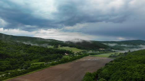 Camiones-Que-Viajan-Con-Niebla-Baja-Y-Nubes-En-Una-Montaña-Verde-Bajo-Un-Cielo-Oscuro---Plano-Aéreo-Amplio