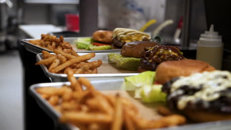 Chef-plates-handful-of-french-fries-on-trio-of-metal-trays-with-burgers-in-restaurant-kitchen,-slow-motion-slider-4K