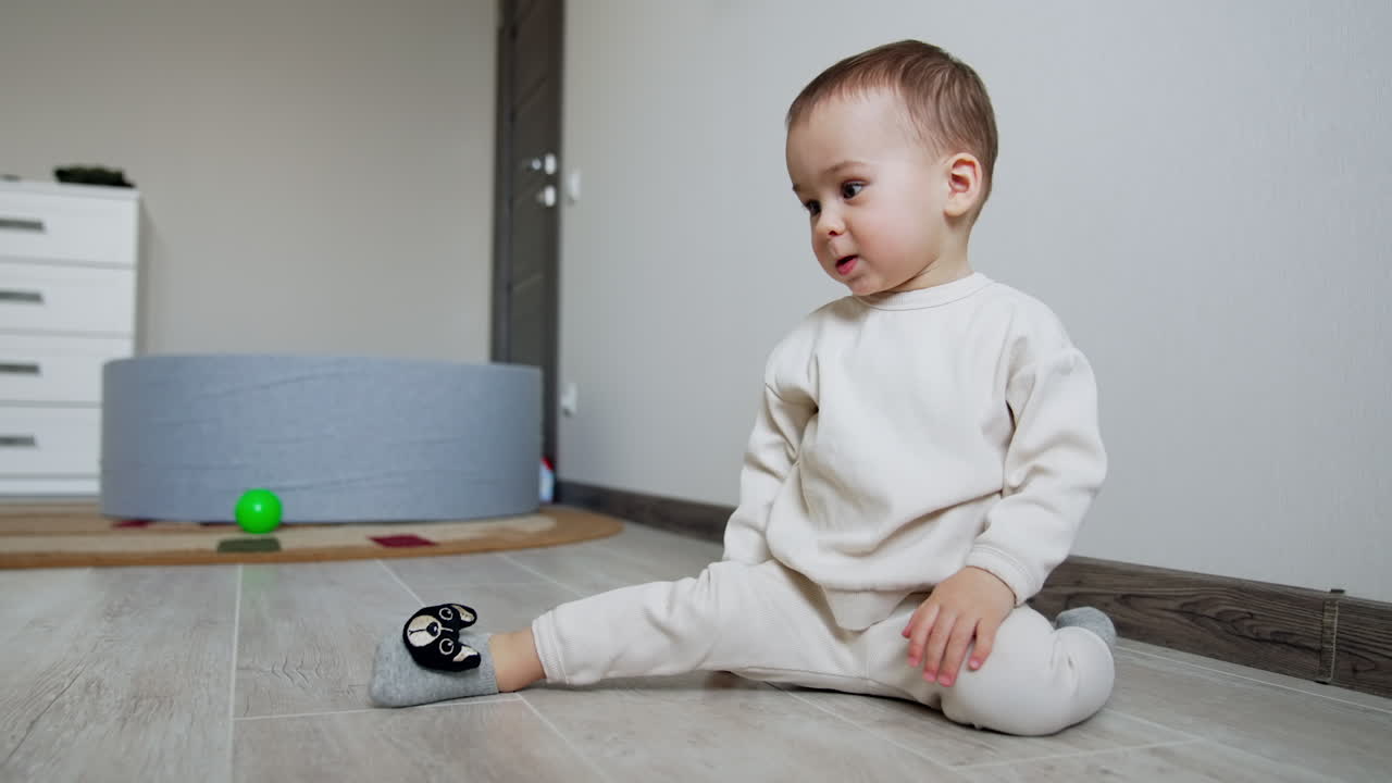 Smiling kid in white sport suit sits on the floor. Baby thinks for a moment and then crawls away.
