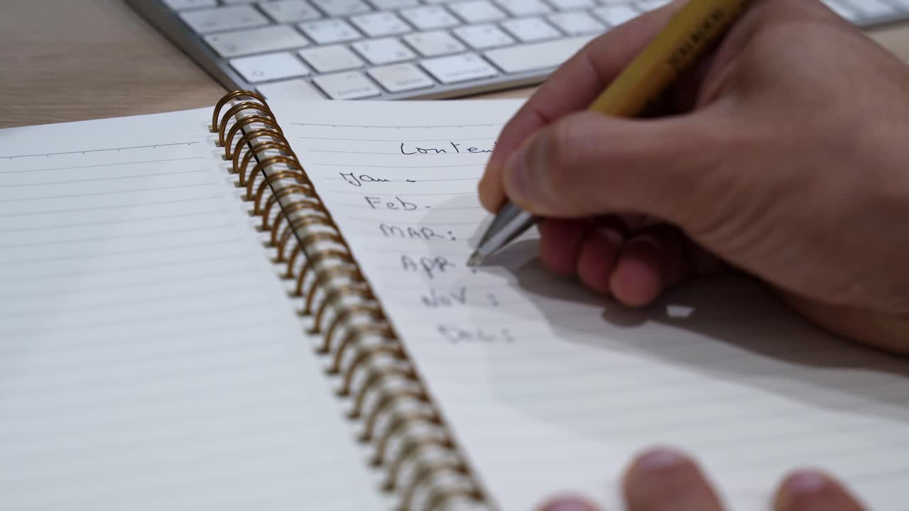 a hand writing a monthly planning list in a gold ring spiral notebook on a wooden desk with an Apple keyboard in the background, ideal for business and marketing content