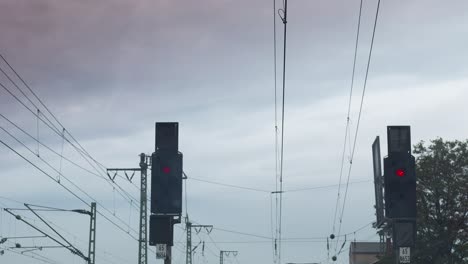 Under-a-gloomy-cloudy-sky,-a-railway-crossing-comes-to-life-with-two-traffic-lights,-one-red-and-the-other-yellow,-surrounded-by-crisscrossing-overhead-wires,-with-trees-and-buildings-in-the-backdrop