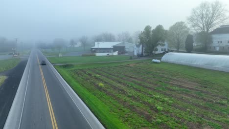 Car-drives-on-road-along-yard-with-greenhouse-and-white-house,-aerial