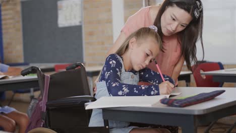 maestra diversa que enseña a una escolar en silla de ruedas escribiendo en el aula de la escuela primaria