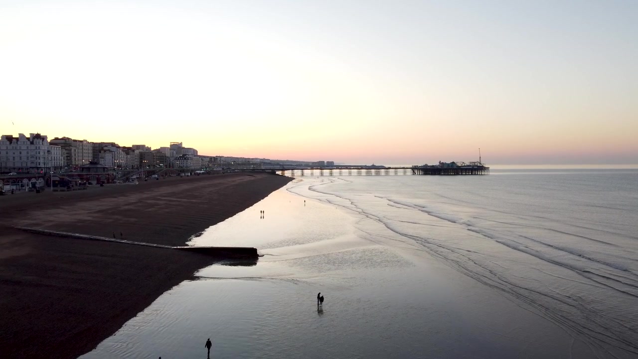 Drone shot running close to the ground along the coastline of Brighton revealing people walking along the beach at low tide whilst the sun rises