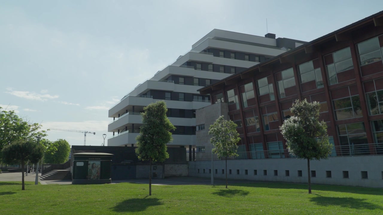 Plano contrapicado de un edificio contemporáneo de uso mixto con fachada de cristal y exterior de ladrillo, césped verde en primer plano, Gijón, España