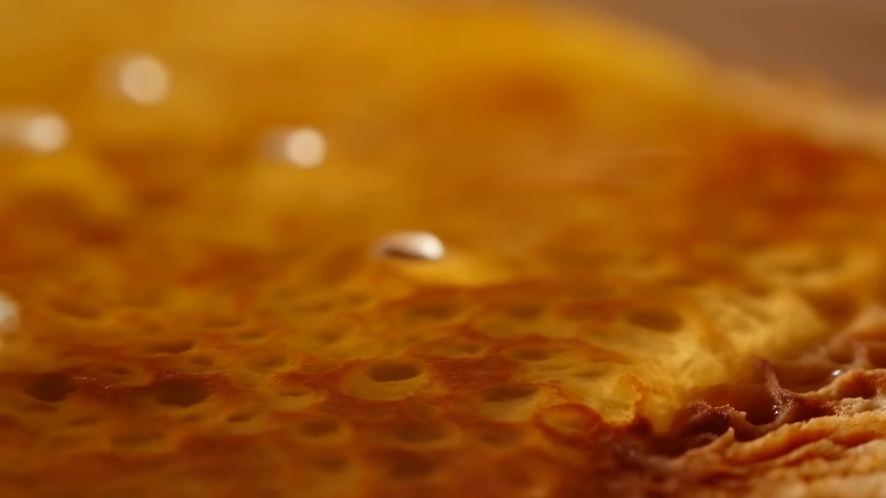 Primo piano estremo in movimento che mostra una superficie alimentare dorata e porosa con luce e messa a fuoco variabili, rivelando cavità simili a un nido d'ape, lucentezza caramellata, cristalli di sale e una texture tattile calda.