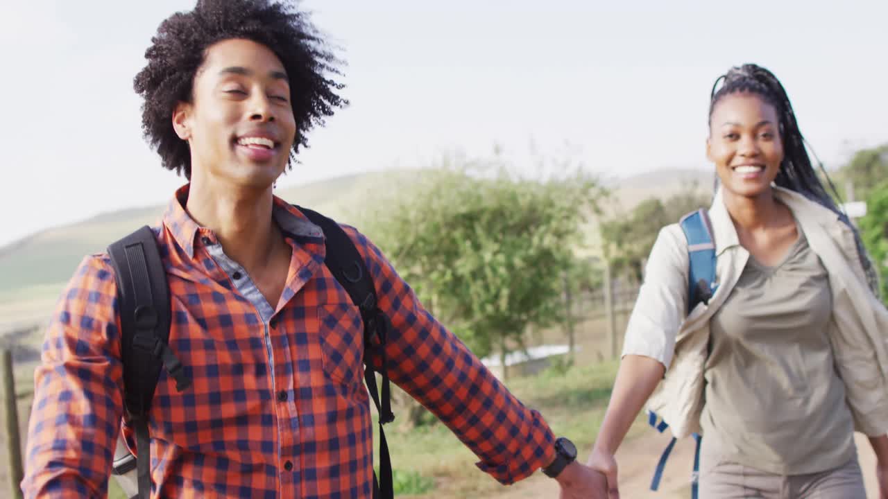 Happy african american couple with backpacks, hiking together on sunny day, slow motion