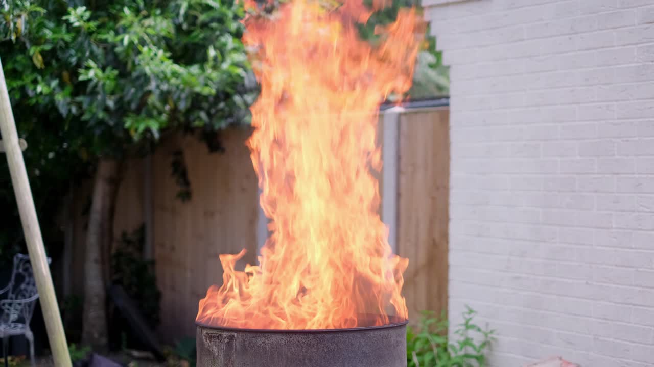 Slow motion footage of a large fire flame coming out of a bonfire drum in garden. The flames flap in the gentle breeze with intense yellow and orange colour. The fire holds the centre of the frame.