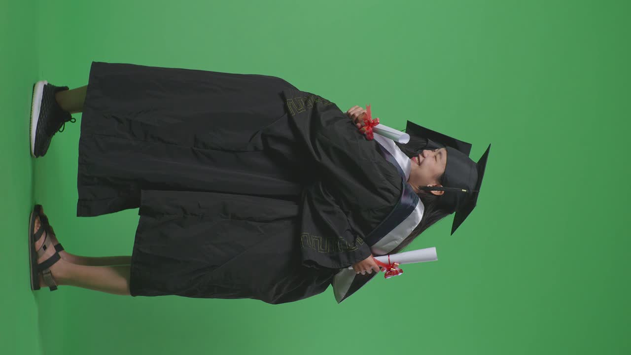 Studentesse asiatiche laureate a figura intera con tocchi e toghe, con i diplomi, che sorridono e si abbracciano su sfondo green screen in studio