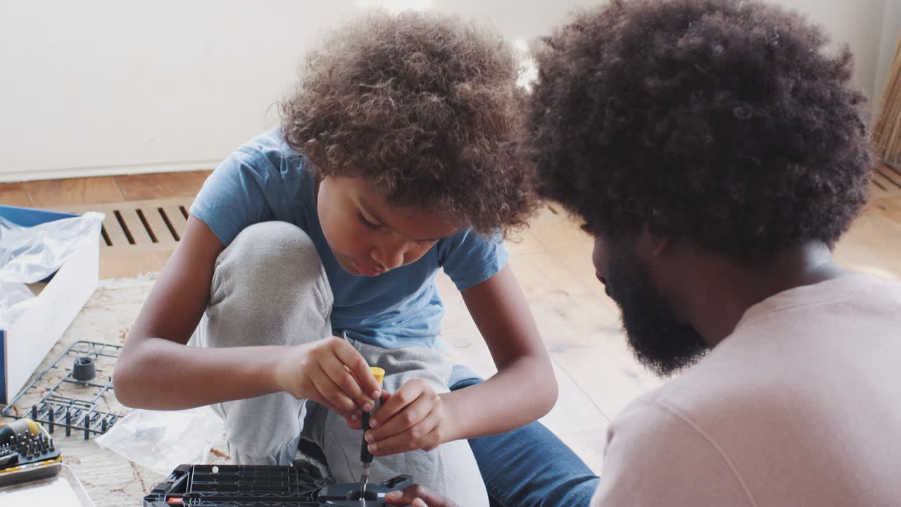 vista sobre el hombro de un niño preadolescente sentado en el suelo de la sala de estar con su padre, usando un destornillador para montar un kit de juguete, de cerca