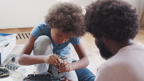 Over shoulder view of pre teen boy sitting on the floor in the living room with his father, using a screw driver to assemble a kit toy, close up