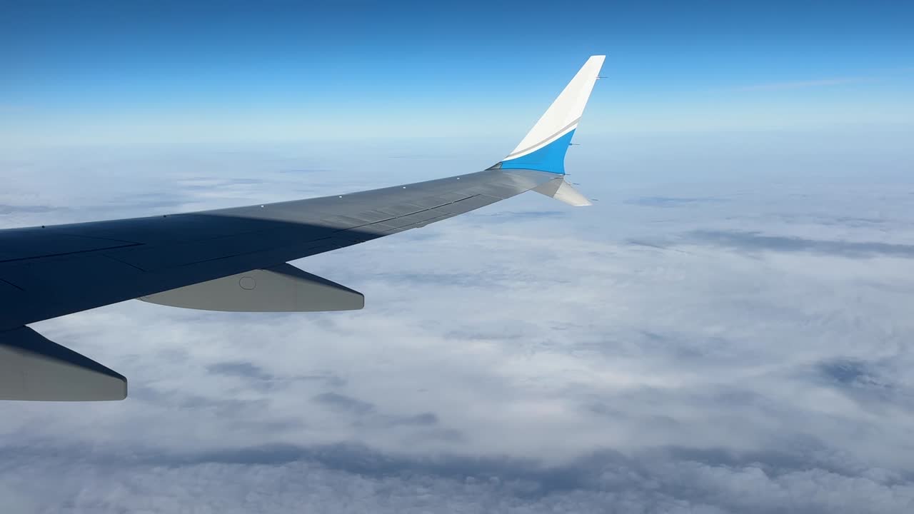 Passasjervinduutsikt av en kommersiell Boeing 737 Max-vinge over spredte skyer i en klar, blå himmel i marsjhøyde, over skyer