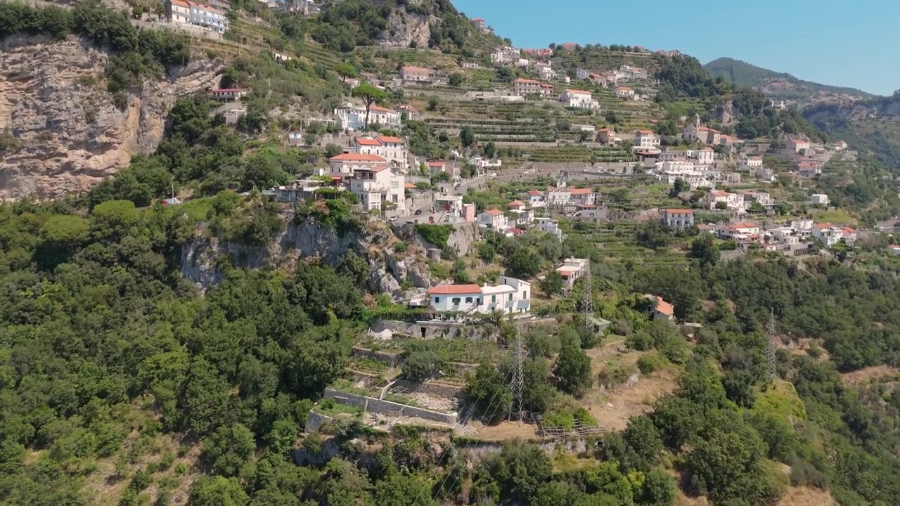 A drone captures a hillside village in Italy with homes built on slopes. The area has terraced fields and greenery, showing the landscape and architecture.