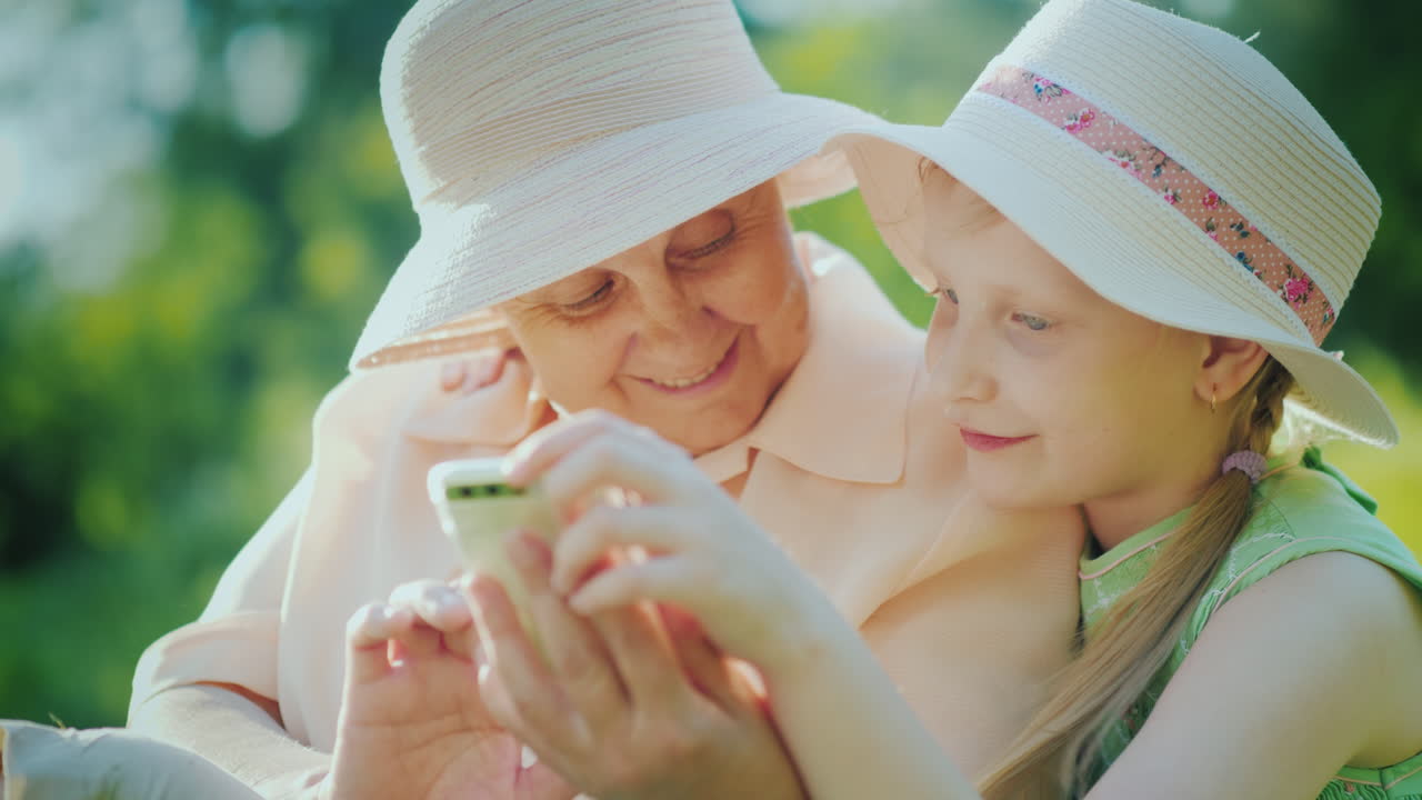 할머니와 손녀는 스마트폰을 함께 사용하여 여름날 4k에 공원에서 휴식을 취합니다.