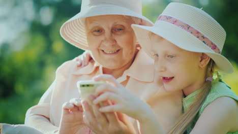 grandmother and granddaughter use the smartphone together have a rest in the park on a summer day 4k