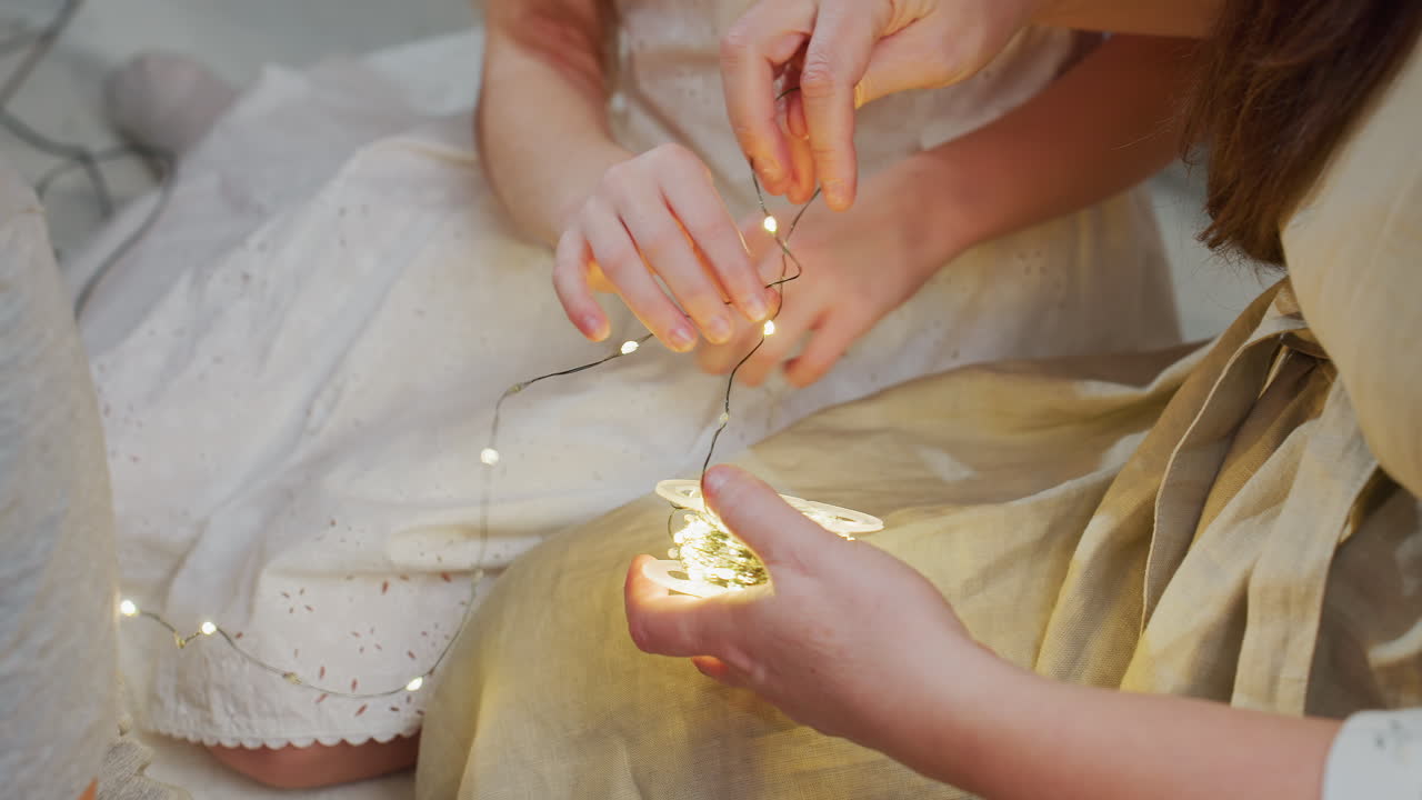 Close-up shot of mother and daughter arranging Christmas lights together, smiling and enjoying the holiday spirit, the mother, in a grey gown, and daughter in a white gown add festive cheer