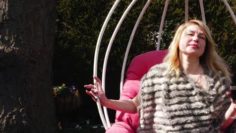 Attractive-woman-enjoys-glass-of-wine-in-garden-chair-swing-wide-zoom-slow-motion