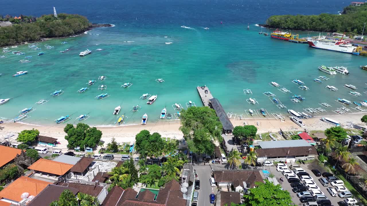 Captivating drone video showcases Padang Bai's vibrant port with turquoise waters and moored boats. Lush greenery and clear blue skies create a picturesque, serene atmosphere