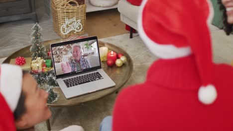 Familia-Diversa-Con-Sombreros-De-Santa-Usando-Una-Computadora-Portátil-Para-Una-Videollamada-Navideña-Con-Un-Hombre-Feliz-En-La-Pantalla