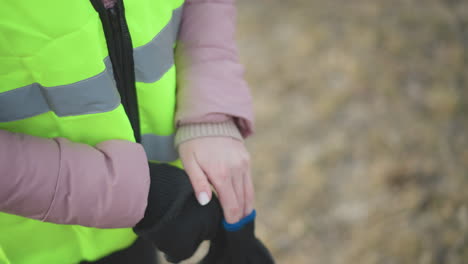 close-up of person in bright high-visibility neon yellow safety vest with reflective stripes and pink padded jacket putting on black knitted gloves outdoors, preparing for work in cold winter conditions