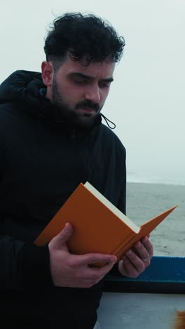 man carefully reads a book while relaxing by the sea in winter