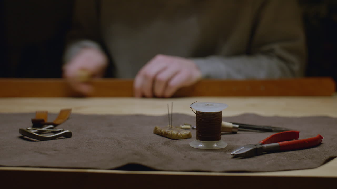 A set of leatherworking tools including thread, needles, pliers, and a leather strap placed on a workbench, with artisan hands working in the background.