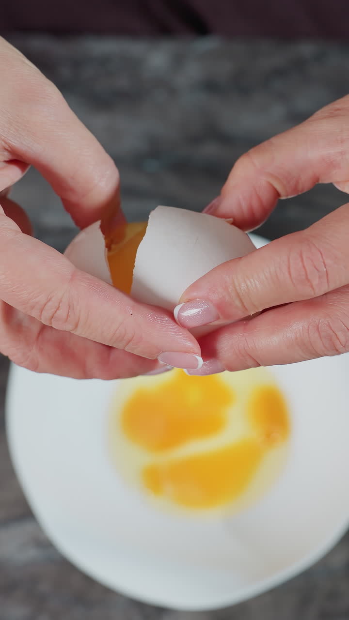 Aerial view of chef's hands adorned with a bracelet cracking an egg into a white bowl, egg yolks are visible inside the bowl, with a cup of sugar nearby on a dark kitchen countertop