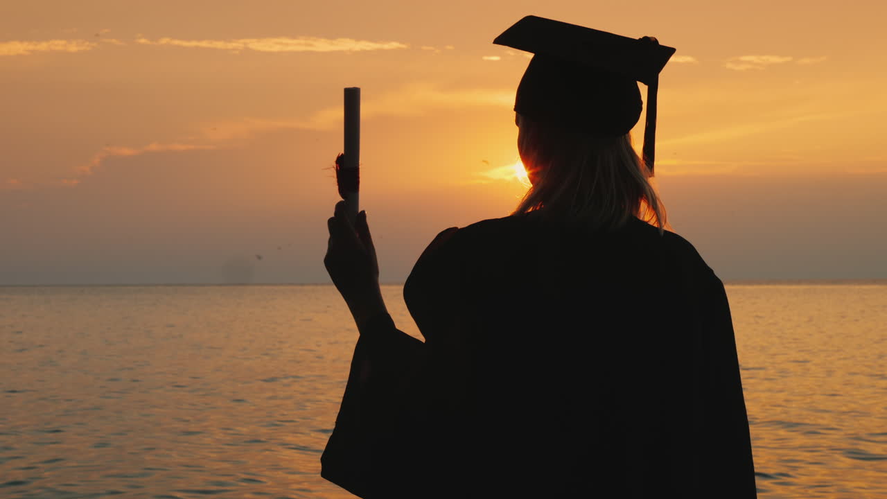 un soltero con un diploma en la mano y una gorra de graduado mira el amanecer sobre el mar