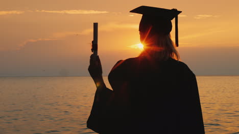 a bachelor with a diploma in hand and a cap of a graduate looks at the sunrise over the sea